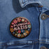 Round pinback metal button featuring an embroidered-style design on a black background. The center showcases bold, stitched-textured lettering in red, blue, and white that reads "PUNCH NAZIS." Surrounding the text is an intricate, folk-inspired floral pattern with multicolored flowers in shades of pink, orange, blue, and white, arranged symmetrically. A decorative zigzag border frames the edge of the button. The backside is silver metal with a secure pin closure.